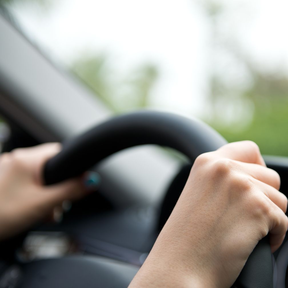 Close-up of a person’s hands gripping a steering wheel while driving a car, with a blurred green background visible through the window.