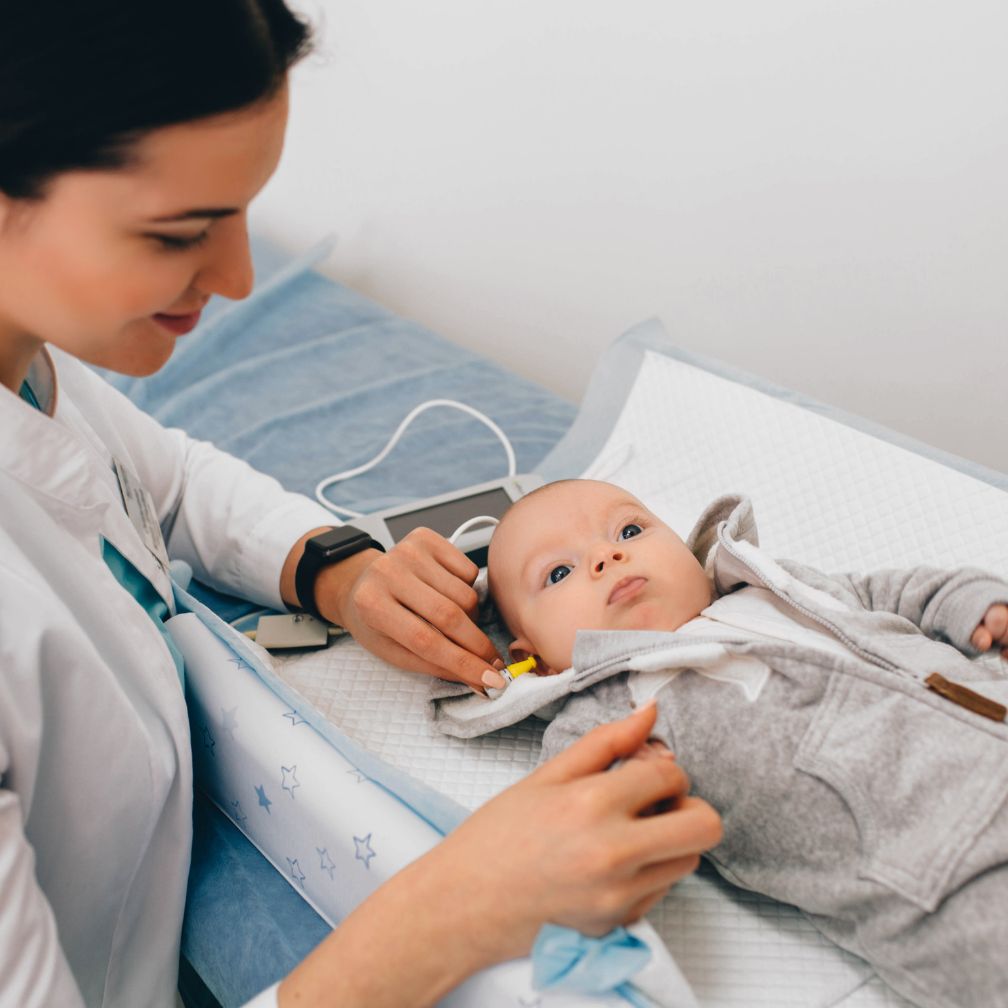 A healthcare professional tests a babys hearing using a small device while the baby lies on an exam table, dressed in a gray outfit.