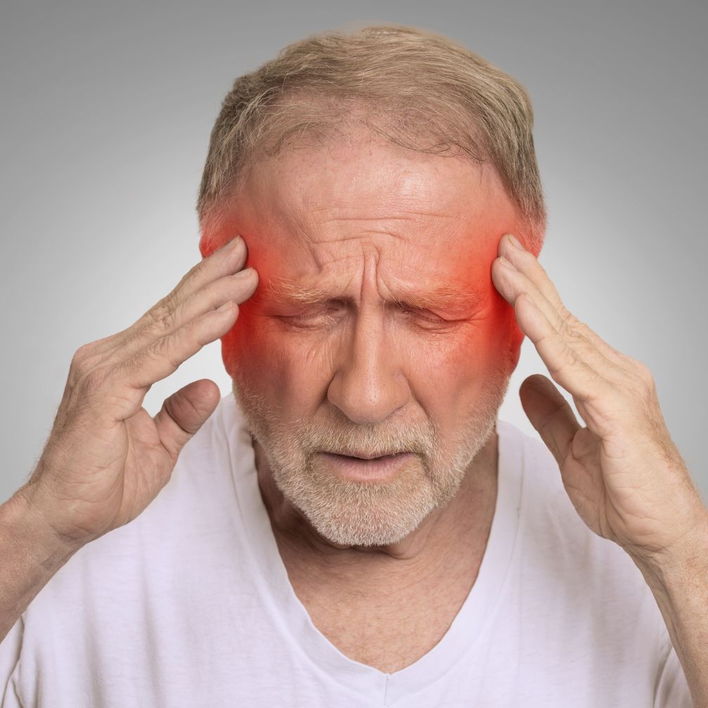 An older man with short hair holds his head with both hands, eyes closed, appearing in pain. Red highlights on his temples suggest a headache or migraine.