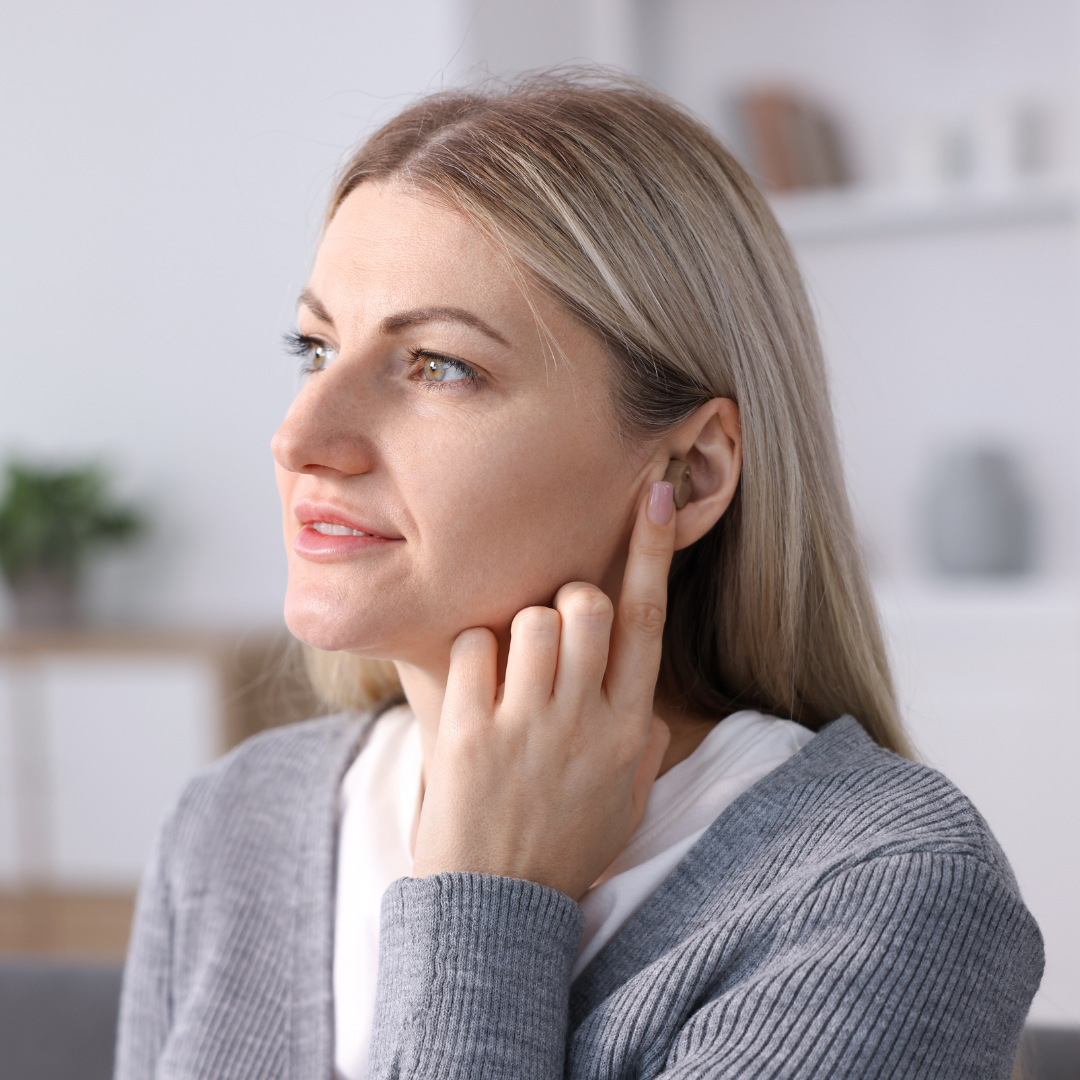 A woman with blonde hair and a gray sweater touches her ear with her finger, appearing thoughtful, in a brightly lit room with blurred shelves and plants in the background.