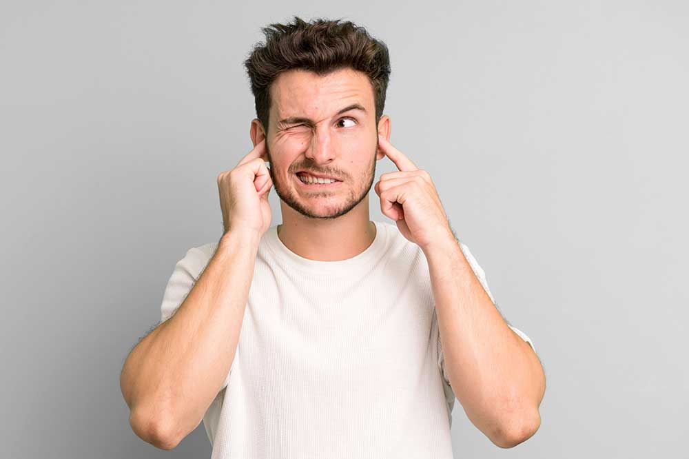Young man grimacing and plugging his ears with his fingers, as when one hearing aid sounds off, from Calvert Hearing Care in North Texas